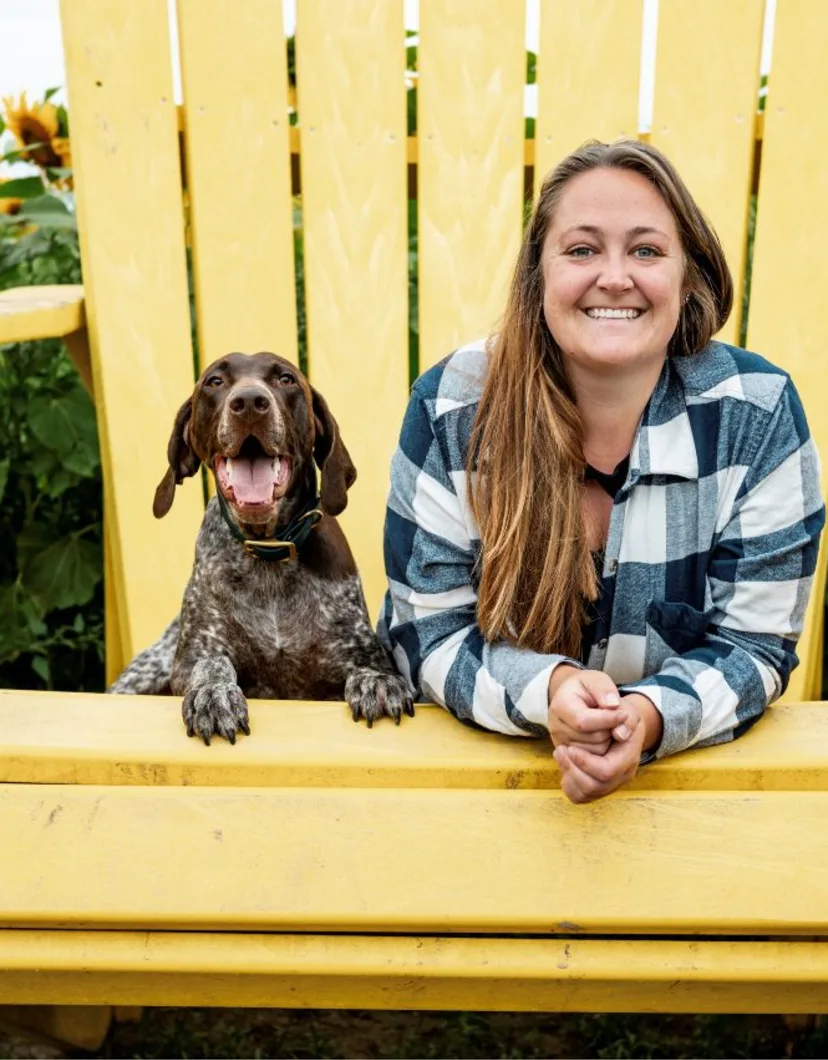 Nicole in a plaid shirt laying on a giant yellow lawn chair with a smiling dog.